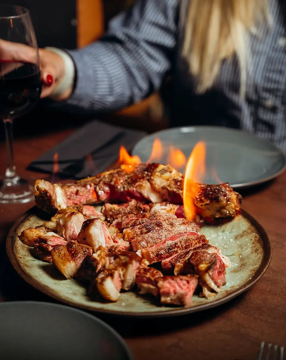 Um prato de carne a flamejar e uma mão com um copo de vinho tinto
